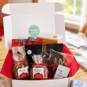 May include: A white gift box filled with assorted snacks including a red bag of cherry berry mix, a red bag of sour cherry gummy bears, a black bag of pretzels, a box of baked goods, and a small white card with the text Just for you.