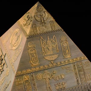 Può includere: Una piramide dorata con intricate incisioni di geroglifici e divinità egizie. La piramide è realizzata in un materiale testurizzato che ricorda il legno.