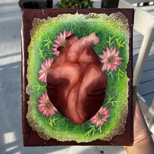 May include: A painting depicting a human heart surrounded by pink flowers on a green background. The heart is painted realistically with a red and brown colour scheme. The flowers are painted in a stylised manner with pink petals and green leaves. The painting is framed in a brown frame.