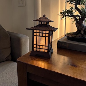 May include: A brown pagoda-style lantern with a warm, glowing interior sits on a wooden table. The lantern has a multi-tiered roof and a grid-like window design. A bonsai tree in a black pot rests on a book nearby.