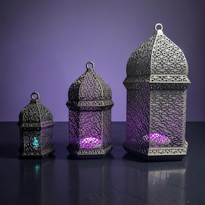 May include: Three metallic lanterns of varying heights featuring intricate geometric cutouts. Each lantern has a domed top with a small hanging loop and contains a glowing light source, emitting purple and teal hues through the patterned metal frame.