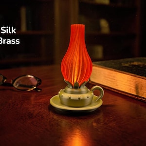Könnte beinhalten: Eine dekorative Lampe mit rotem, geripptem Schirm und messingfarbenem Sockel. Die Lampe steht auf einer kleinen, runden Platte mit Griff. Der Text "Silk Brass" ist sichtbar. Die Lampe steht auf einer Holzoberfläche.