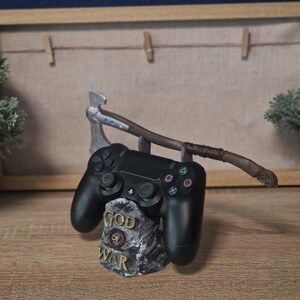 Può includere: Un controller nero per PlayStation 4 è posizionato su un supporto in pietra grigia con la scritta "God of War". Un'ascia argentata con un manico in legno marrone è appoggiata sul supporto.