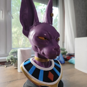 May include: A purple cat-like humanoid bust featuring large pointed ears, yellow eyes, and a gold collar. The figure wears a decorative neck piece with black, blue, and white segments, a gold border, and a red diamond-shaped emblem on a black base.