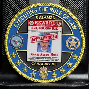 May include: A commemorative coin with a blue and gold border, featuring text that reads "EXECUTING THE RULE OF LAW." The coin includes a "REWARD" notice for $50,000,000 USD and a photo of Nicolás Maduro Moros. The coin also has the text "APPREHENDED."