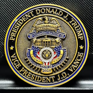 Pu&ograve; includere: Una moneta commemorativa dorata con un anello esterno nero e blu. La moneta presenta un'aquila, la Casa Bianca e il testo "PRESIDENT DONALD J. TRUMP IN COMMEMORATION OF VICE PRESIDENT J.D. VANCE 2025".