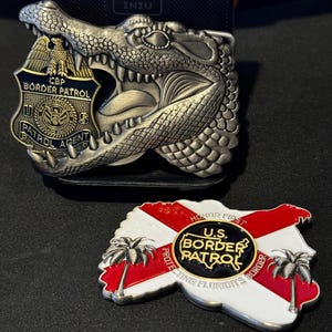 May include: Two metal belt buckles. One is a silver alligator head with a CBP Border Patrol badge. The other is a Florida-shaped badge with the text "U.S. Border Patrol" and "Protecting Florida's Border."
