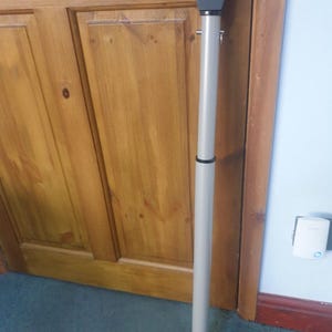 May include: A grey door security bar with a black base and a silver top. The adjustable bar is designed to prevent forced entry. It is positioned against a wooden door.