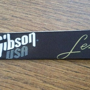 May include: Black key chain with a silver metal ring. The key chain has white text that says "Gibson USA" and gold text that says "Les Paul".