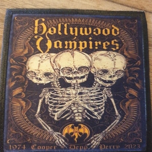 May include: A rectangular patch with a dark blue border features the words "Hollywood Vampires" in gold lettering above a skeletal figure with three skulls. The design includes a bat emblem and the years 1974 and 2023.