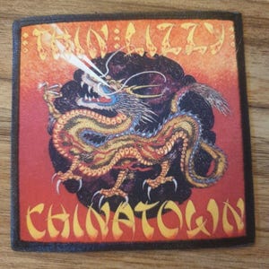 May include: Square patch with a golden dragon illustration, detailed with red and blue. The patch features the words "Thin Lizzy" and "Chinatown" in yellow, set against a red and black background, reminiscent of a vintage design.