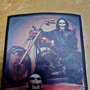 May include: A black and white patch featuring a man in a black suit and red bow tie. He is holding a motorcycle with a skull mask on the ground. The man is wearing a mask with a red spiral design.