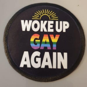 May include: A round black patch with a white border and the text "Woke Up Gay Again" in white. The word "Gay" is in rainbow colours.