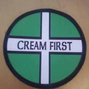 May include: A round, green and white patch with a black border. The patch has a white cross in the centre and the words "CREAM FIRST" written in black letters.