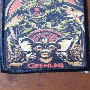 May include: A black and white embroidered patch featuring a colourful image of two Gremlins. The top Gremlin is green and has a menacing expression. The bottom Gremlin is brown and has a mischievous expression. The patch has the word "GREMLINS" printed in red at the bottom.