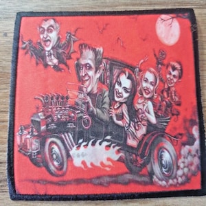May include: A square patch with a red background depicting a cartoon image of the Munsters family in a hot rod car. The image includes Frankenstein, Lily Munster, Herman Munster, and Grandpa Munster. A full moon and bats are in the background.