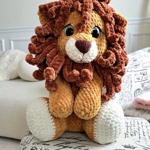 May include: A handmade crochet lion plush toy. The lion features a light orange body, a brown mane, and white paws. It has large black eyes, a white muzzle, and a brown nose. The plush is sitting on a patterned ottoman.
