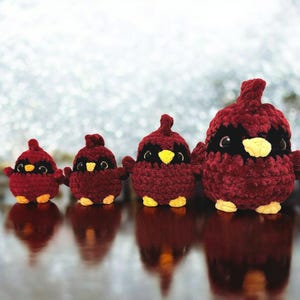 Op de afbeelding: Vier pluche kardinaalvogels in verschillende maten, allemaal gemaakt van rood garen met zwarte ogen en een gele snavel. De vogels zijn in een rij gerangschikt van de kleinste tot de grootste.