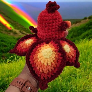 Op de afbeelding: Een handgemaakte, rode gehaakte octopus met grote, bloembladachtige aanhangsels in tinten oranje en geel. De octopus heeft een getextureerd uiterlijk en een afgeronde kop. De achtergrond toont een regenboog en een grasrijke heuvel.