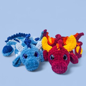 May include: Two plush crocheted dragons in a light blue setting. One dragon is blue with white accents, the other is red, orange, and yellow. Both have large, round, black eyes and are lying down.
