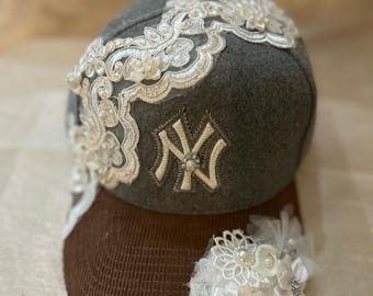 Peut inclure: Une casquette de baseball grise avec un dos en mesh marron. La casquette est ornée d'une bordure en dentelle blanche et d'un logo "NY" brodé en blanc. La casquette est décorée de fleurs en dentelle blanche et de perles.