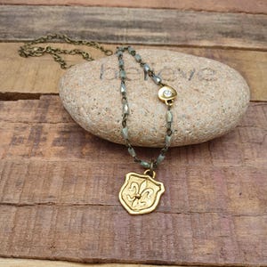 Puede incluir: Un collar dorado con un colgante de flor de lis. El collar presenta una cadena con pequeñas cuentas grises y un pequeño dije redondo con una piedra transparente. El colgante tiene forma de escudo con un diseño detallado de flor de lis.