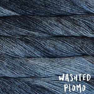 May include: Close-up of a skein of Washted Plomo yarn. The yarn is a blend of dark and light blues, creating a textured appearance. The text "WASHTED PLOMO" is written in white script across the bottom of the image.
