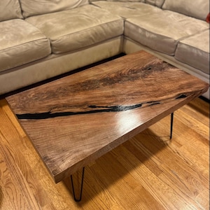 May include: A rectangular coffee table with a natural wood grain finish and a black resin inlay. The table has a dark brown color and is supported by four black hairpin legs. The table measures approximately 48 inches long and 24 inches wide.