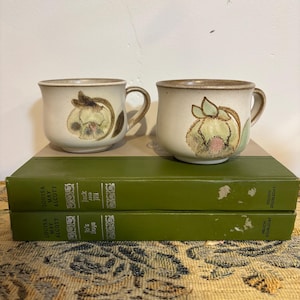 May include: Two ceramic mugs with floral designs in shades of green, brown, and pink. The mugs sit atop two green hardcover books. The mugs have a beige background with a brown rim and handle.