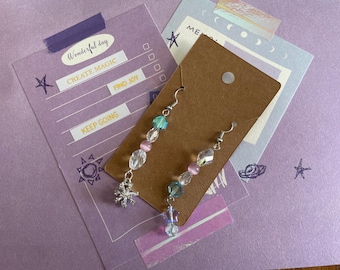 May include: Dangle earrings with clear, pink, and blue beads, and a silver snowflake charm. The earrings are displayed on a brown card against a purple background with the words "Wonderful day", "Create Magic", "Find Joy", and "Keep Going".