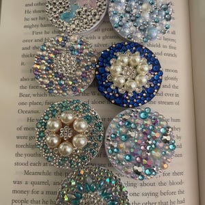 May include: A collection of bedazzled phone grips in various colors and designs. Each grip is round and covered in rhinestones, pearls, and other decorative elements. The grips are arranged on an open book.