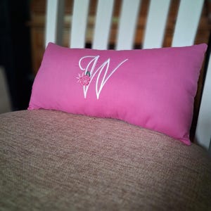 May include: A rectangular pink pillow with a white embroidered letter "W" and a small floral design. The pillow is placed on a brown textured surface, with a white chair back in the background.