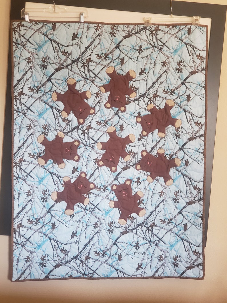 K&ouml;nnte beinhalten: Eine blaue und braune Tarnstoffdecke mit acht braunen Teddyb&auml;r-Applikationen. Die B&auml;ren sind alle in die gleiche Richtung ausgerichtet und haben ein einfaches gesticktes Gesicht.