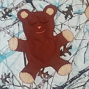 K&ouml;nnte beinhalten: Ein brauner Teddyb&auml;r mit geschlossenen Augen und einer rosa Nase liegt auf einem wei&szlig;en und blauen Tarnstoff. Der Stoff tr&auml;gt den Schriftzug "TrueTimber".