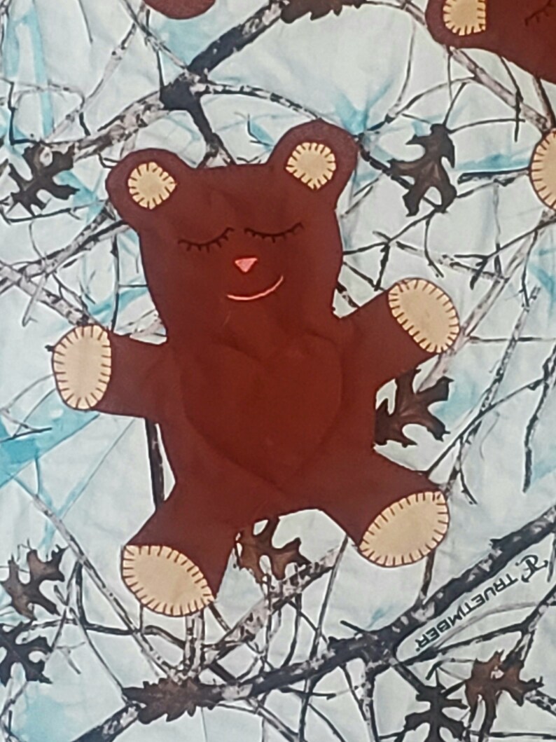 K&ouml;nnte beinhalten: Ein brauner Teddyb&auml;r mit geschlossenen Augen und einer rosa Nase liegt auf einem wei&szlig;en und blauen Tarnstoff. Der Stoff tr&auml;gt den Schriftzug "TrueTimber".