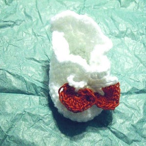 May include: A handmade white crochet baby shoe with two small red bows. The shoe is made of soft yarn and is designed for a newborn. The shoe is sitting on a textured teal surface.