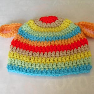 May include: A handmade crochet hat in a rainbow of colors, including blue, yellow, orange, and red. The hat features two orange ear-shaped accents. The hat is likely for a baby or toddler.