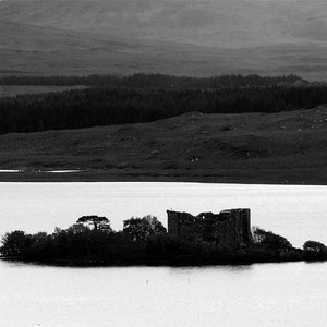 May include: A black and white photograph of a stone castle ruin on a small island in a loch. The castle is surrounded by trees and the loch is calm and still. The background shows a distant mountain range.