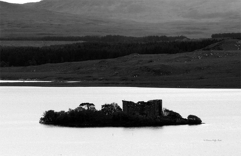 May include: A black and white photograph of a stone castle ruin on a small island in a loch. The castle is surrounded by trees and the loch is calm and still. The background shows a distant mountain range.