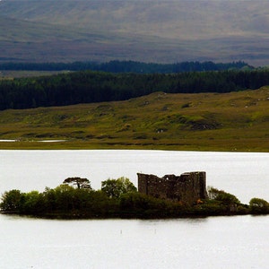 May include: A ruined stone castle on a small island in a lake. The island is covered in green trees and bushes. The lake is calm and still, reflecting the sky and the surrounding hills.