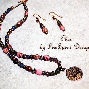 May include: A multi-strand necklace with pink, brown, and iridescent beads, a copper pendant with an intricate design, and matching earrings. The necklace is labeled "Elise by Fire Spirit Designs".