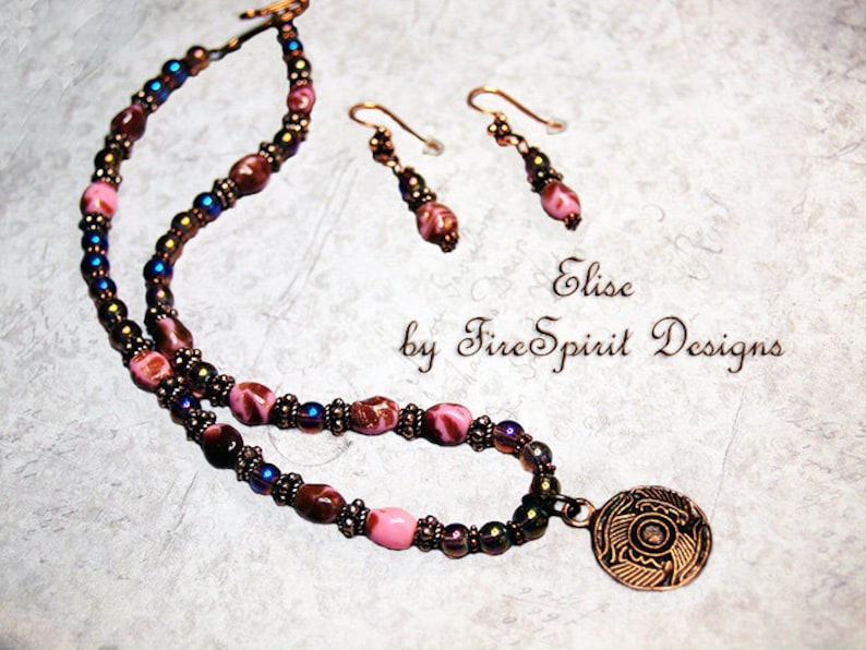 May include: A multi-strand necklace with pink, brown, and iridescent beads, a copper pendant with an intricate design, and matching earrings. The necklace is labeled "Elise by Fire Spirit Designs".