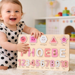 May include: Wooden educational puzzle featuring the name "PHOEBE", numbers 1-10, and shape cutouts. The puzzle has pink and white accents. The shapes include a square, hexagon, triangle, and star. The puzzle is designed for early learning.