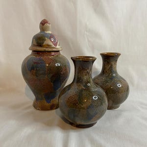 May include: Three ceramic vases with a mottled glaze in shades of brown, blue, and red. The set includes a lidded jar and two smaller vases. The vases have a glossy finish and a classic shape, suitable for home decor.