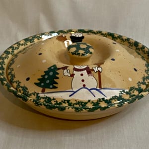 May include: Oval ceramic serving dish with a lid, featuring a snowman design. The lid has a knob and is decorated with a green and white border. The snowman is wearing a scarf and holding a tree. The dish is beige with blue and white accents.