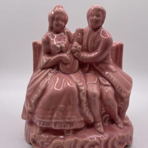 May include: A pink ceramic planter shaped like a couple sitting on a bench. The planter is glazed in a light pink color and has a detailed design.