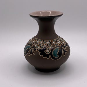 May include: A brown ceramic vase with a decorative floral pattern in teal, black, and white. The vase has a rounded body and a flared neck with a pink interior. The vase is approximately 6 inches tall.