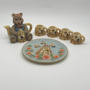 May include: A miniature tea set featuring a teddy bear teapot, four small mugs, and a decorative plate. The teapot is brown with a bear design, and the mugs and plate have bee and floral accents. The plate is light blue with a floral border.