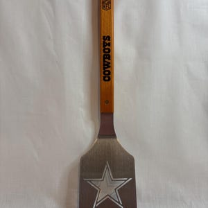 May include: A metal spatula with a wooden handle featuring the NFL logo and the word COWBOYS printed in black. The metal blade is engraved with a large star design, and the top of the handle includes an integrated metal bottle opener.