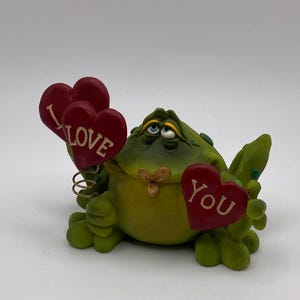 May include: A whimsical frog figurine in shades of green and yellow, holding red heart-shaped signs. One sign reads "I LOVE" and the other says "YOU" in white text. The frog has blue eyes and a small flower detail.